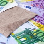Ein hellblauer Geschenkgutschein im Wert von 25 Euro liegt neben einem Sparschwein und ein paar Münzen auf einem Holztisch.