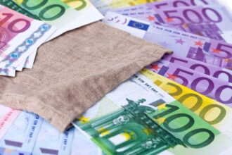 Ein hellblauer Geschenkgutschein im Wert von 25 Euro liegt neben einem Sparschwein und ein paar Münzen auf einem Holztisch.